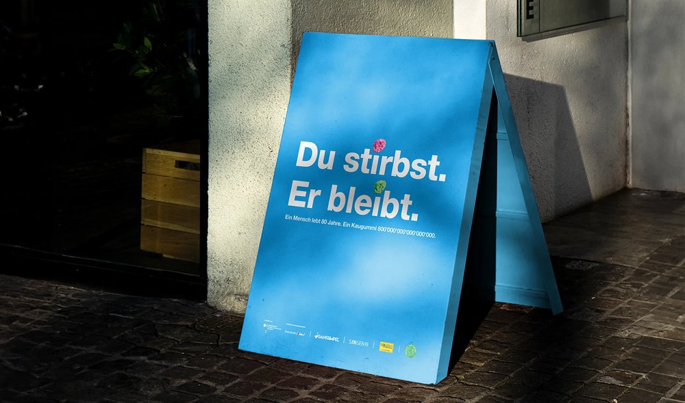 Blaues A-Board-Plakat auf der Straße mit dem Text ‚Du stirbst. Er bleibt.' und einer Erklärung zur Lebensdauer von Kaugummi